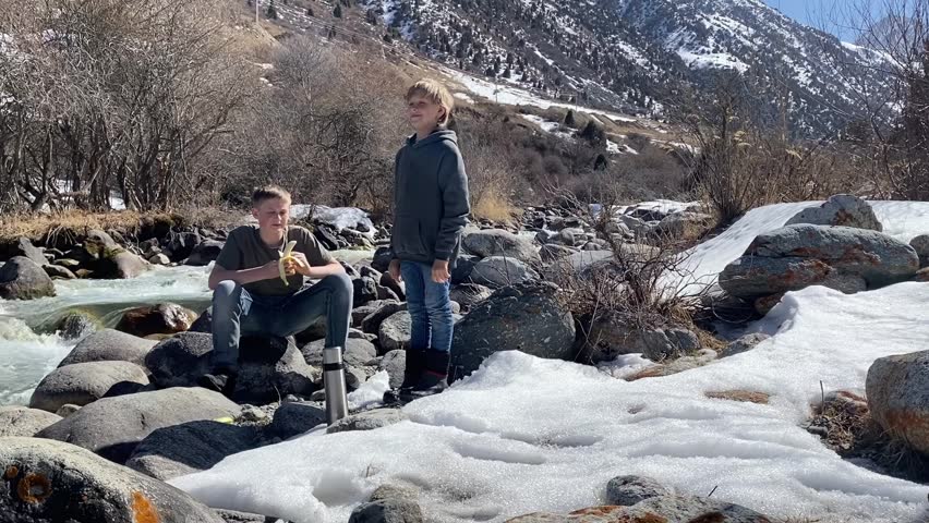 Two boys on rocks by snowy river, sunny day mountain hiking break