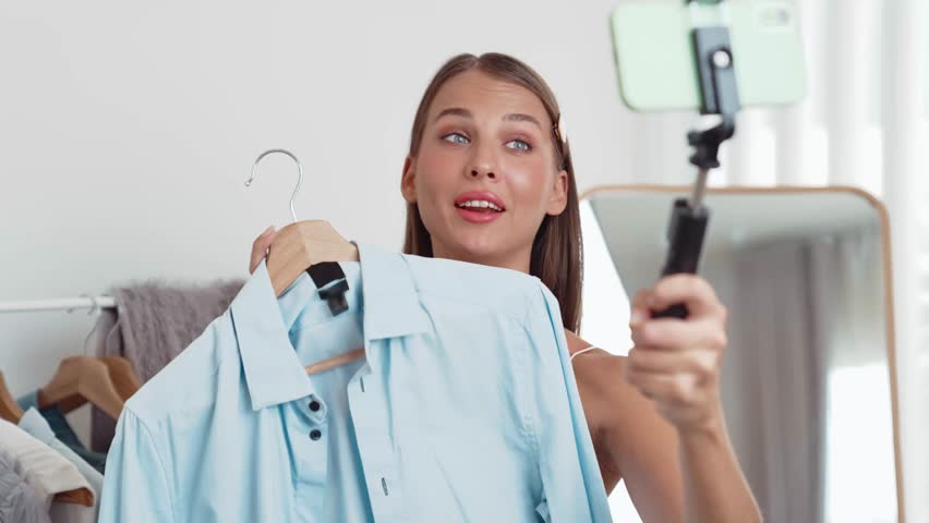 Young social medial content creator woman make fashion video. Blogger smiles to camera using selfie stick and light ring while making persuasive online clothing sell vlog to audience or follower. Adit