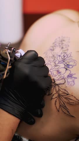 Close-up, in a tattoo salon , a specialist is doing a tattoo on woman's back, black paint floral ornament. a man works in special black gloves, on special equipment.