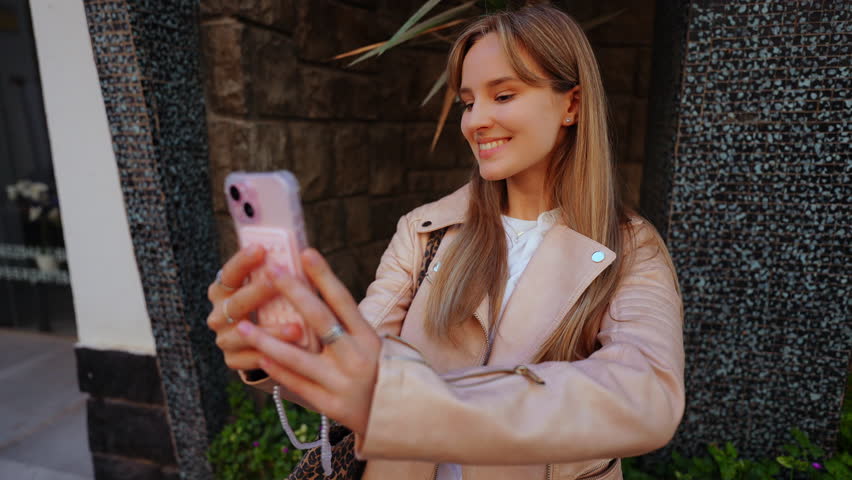 Young woman capturing self portraits while posing dynamically, showcasing trendy lifestyle through smartphone photography in urban outdoor setting