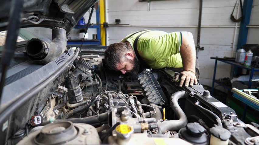 Professional repairman inspecting car engine at service. Adult auto mechanic working under hood of vehicle at garage. Male repairer checking automobile motor at workshop. Transport maintenance