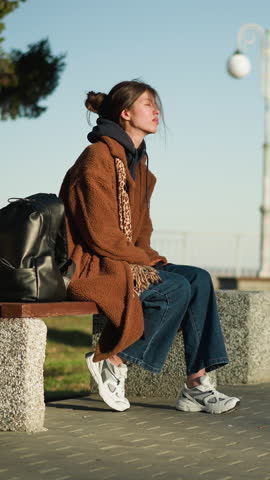 A girl sits alone on a bench in an urban park, appearing somber and lost in thought with her eyes closed. She wears a brown coat, blue jeans, and white shoes, with a black backpack beside her