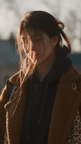 A girl wearing a brown coat and a hoodie is walking faintly, with an expression of weariness and contemplation. Her hair is gently tousled by the wind, moment of quiet reflection in an urban setting