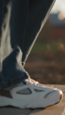 A close-up shot of a person wearing jeans and white shoes, walking slowly on a paved path in a park. The warm, soft light and blurred background