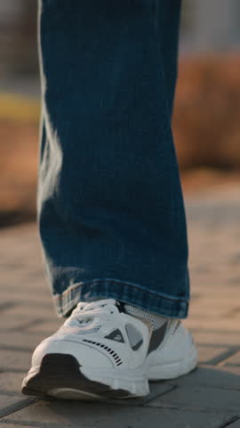 A close-up image of a person wearing jeans and white shoes walking slowly on a paved path in a park. a sense of calmness mixed with sadness, as the deliberate, slow steps reflect a subdued mood