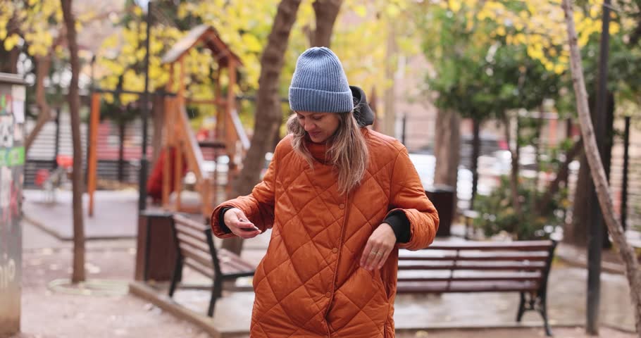 Young woman takes cash from pocket standing at playground in park. Female in orange jacket and knitted hat counts money to make purchase slow motion