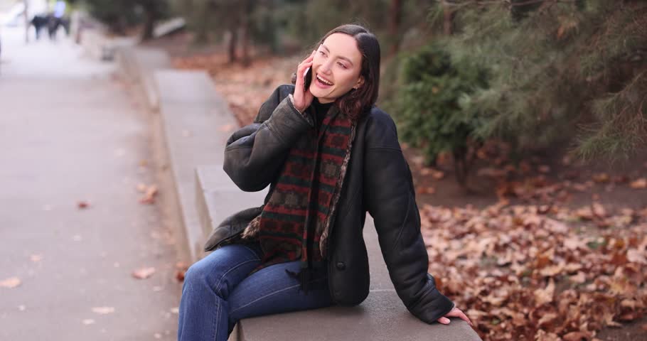 Cheerful young woman in stylish jacket talks on phone sitting in park. Smiling lady in fashionable leather attire enjoys conversation on autumn day slow motion