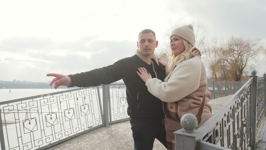 A happy couple in love is walking together on the street in a winter park near a lake. A man and a woman are holding hands, hugging and kissing.