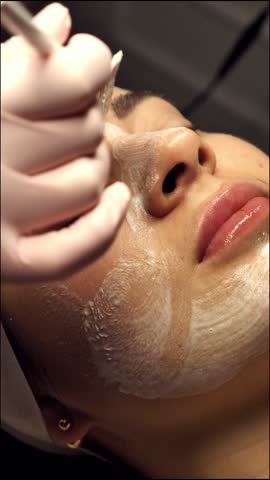 A beautician applies a white cleansing peeling mask to a female face close-up, vertically. A woman at a relaxing spa treatment enjoys