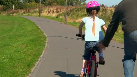Little girl learning to ride her bicycle with help from her dad - dolly Stock Video