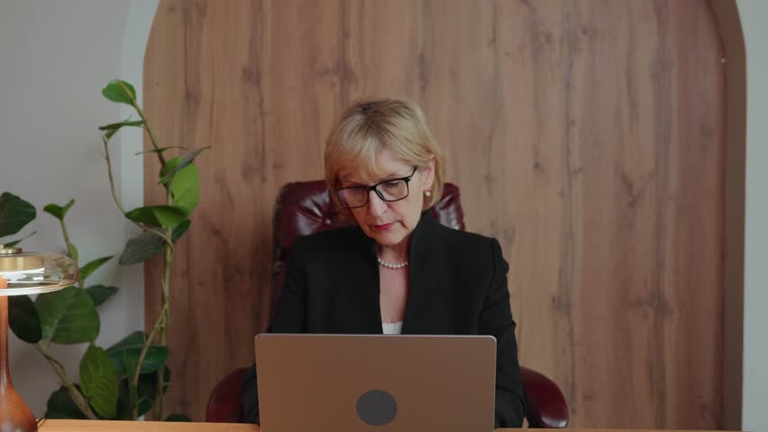 A slim and focused 60-year-old blonde woman works at her laptop. The camera slowly zooms out from a close-up to a medium shot, revealing a calm and professional remote work environment. 4k footage