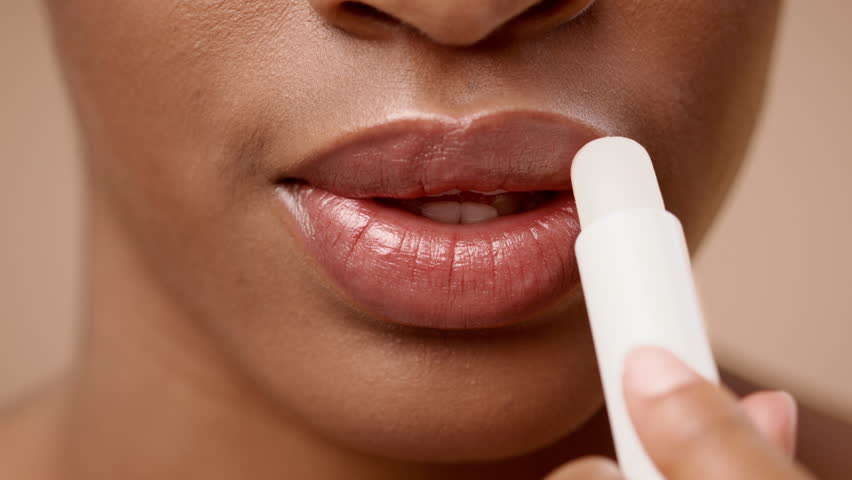 A young African American woman with short hair gently applies lip balm to her lips, focused on enhancing their hydration. The soft background complements her natural beauty.