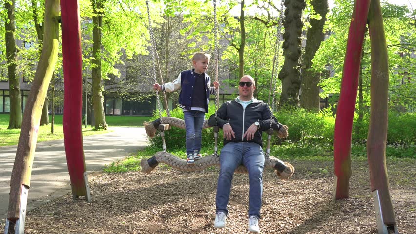Father and child happily swinging together on a large swing in a park. Joyful expressions and relaxed atmosphere highlight the fun, family bonding in a lush green environment.