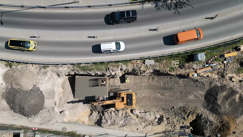 Aerial view of a road construction site with excavators, trucks, gravel piles, and nearby traffic on a curved road
