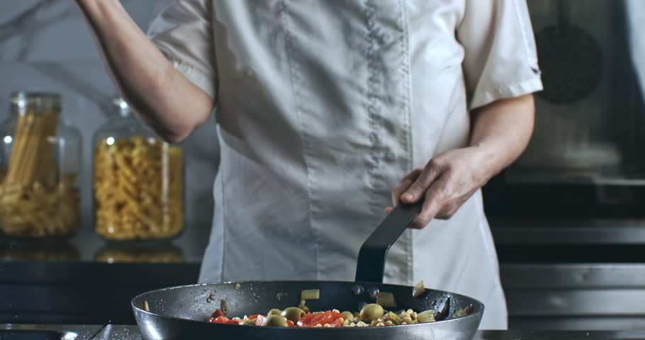 Super Slow Motion of Chef Sprinkling Salt Over Mediterranean Pasta Dish in Pan. Final Seasoning Touch in Professional Kitchen with Fresh Vegetables and Olives on a Stove in a Culinary Environment