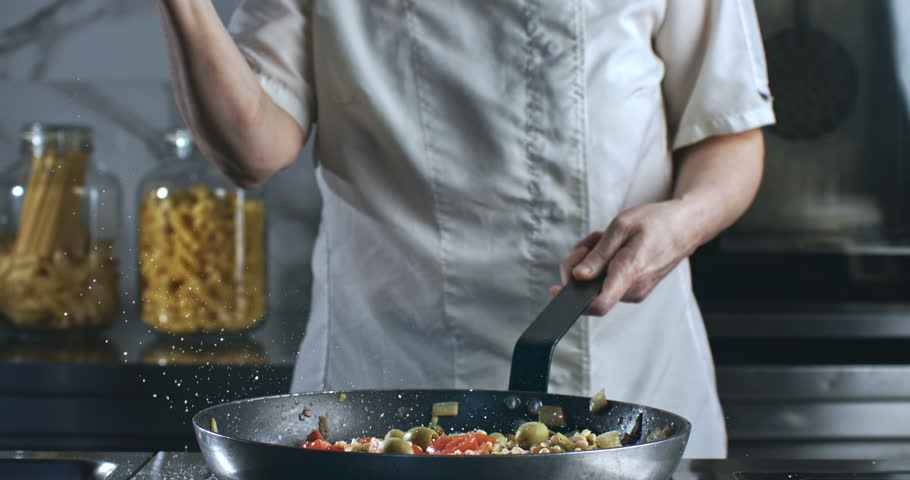 Super Slow Motion of Chef Sprinkling Salt Over Mediterranean Pasta Dish in Pan. Final Seasoning Touch in Professional Kitchen with Fresh Vegetables and Olives on a Stove in a Culinary Environment