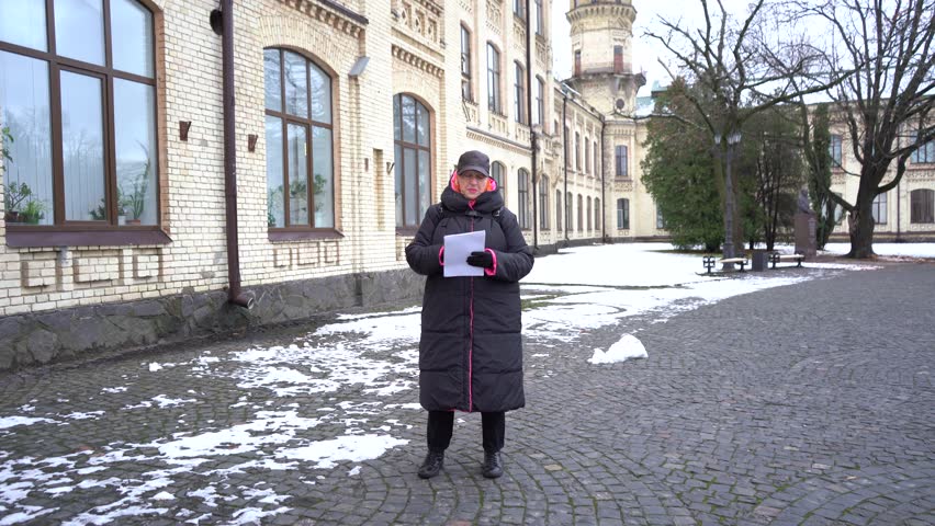 Elderly female inspector inspects an old house from the outside in winter and makes notes in a notebook about its condition. Snow outdoor.