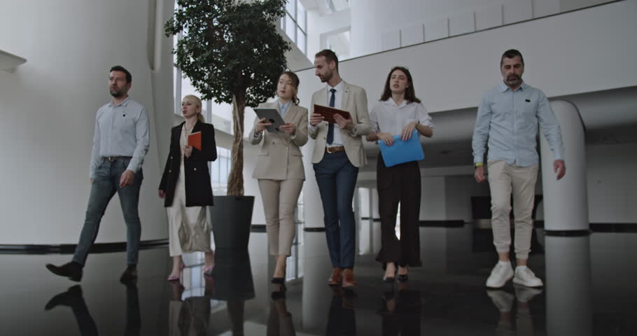 A business team walks and talks in a modern open space office building while reviewing notes, files and tablets.