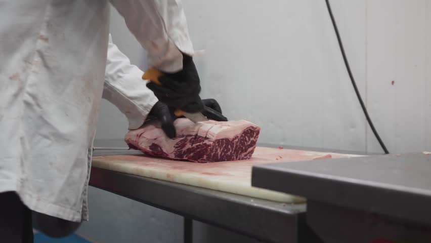 A butcher slices a large, marbled slab of raw beef in an industrial meat processing facility.