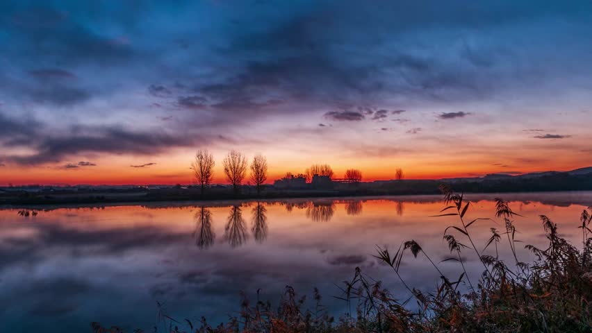 Timelapse of vibrant sunset reflecting on a tranquil lake with silhouetted trees