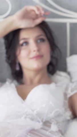 pretty bride with manicure and brilliant earrings lies on bed and looks at long white wedding dress slow motion