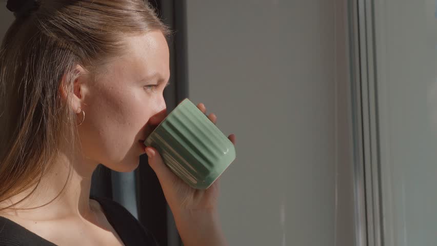Young serene woman drinking coffee or tea from green mug and looking out the window enjoying sunrise and taking a coffee break.