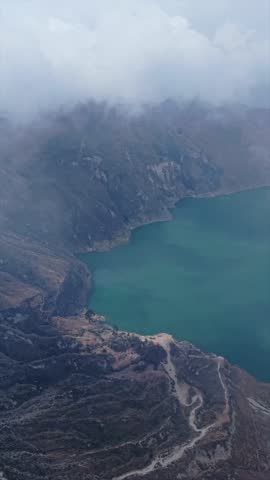 Vertical aerial panorama of Quilotoa volcanic crater, misty clouds sweeping over turquoise waters, Ecuador landscape with changing atmospheric conditions