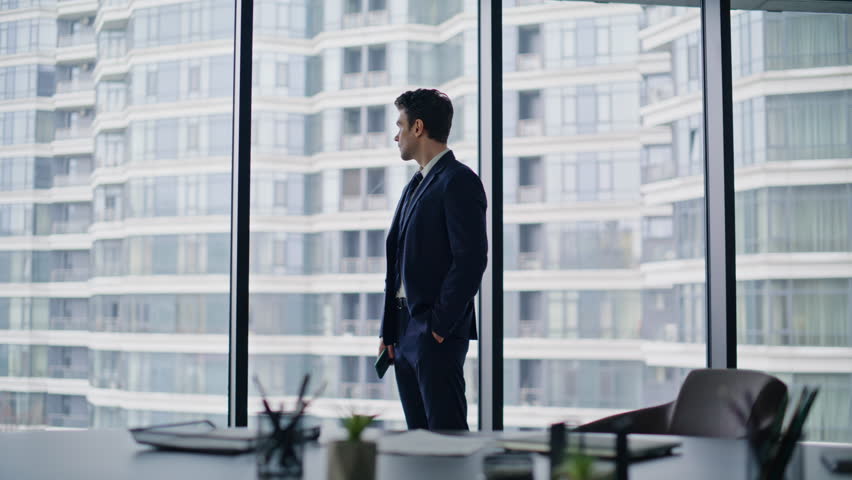 Confident leader standing panoramic office window thinking on business success. Elegant businessman looking on urban high-rise buildings. Handsome man executive holding smartphone pondering project.