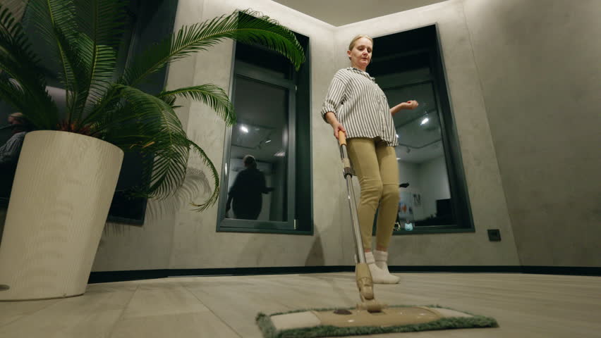Middle-aged woman playfully dancing while mopping indoors, turning a cleaning routine into a fun and energetic moment in a modern room. High quality 4k footage