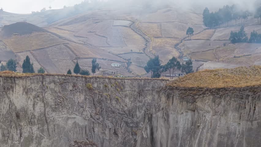 Rural landscape featuring farmland bordering steep canyon rim in Ecuadorian highlands, misty peaks and rising smoke creating geographical panorama