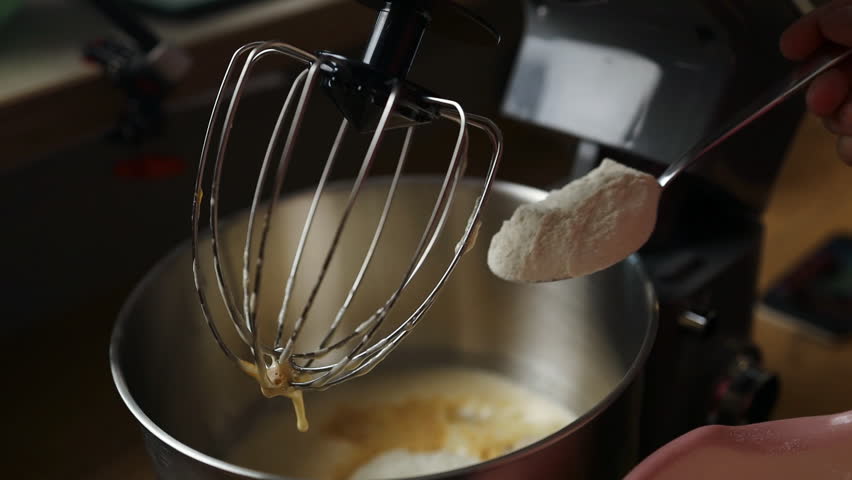 Close-up view of flour being added by spoon into a stand mixer bowl with batter. A step-by-step moment in baking preparation, ideal for cooking tutorials, food blogs, and artisan recipe content.