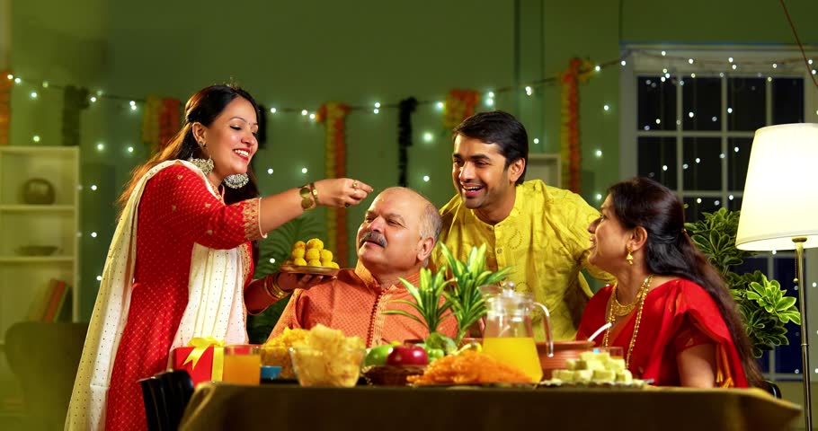 Eating laddoo or laddu and smiling, Indian family of four with senior parents young adult children celebrates Diwali festival at home, seated at decorated dining table looking at camera joyfully