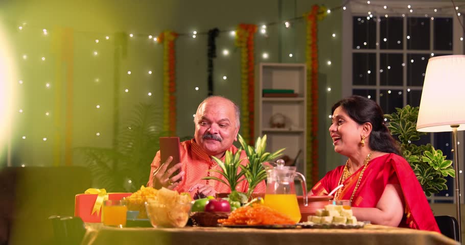 Wishing diwali on video call, Indian family of four with senior parents and young adult children celebrating Hindu festival at home, smiling and talking to relatives at decorated dining table indoors