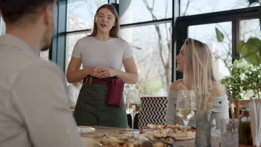 Professional waitress communicating with customers at restaurant table. Friendly service staff explaining menu while guests enjoying wine and food in modern venue