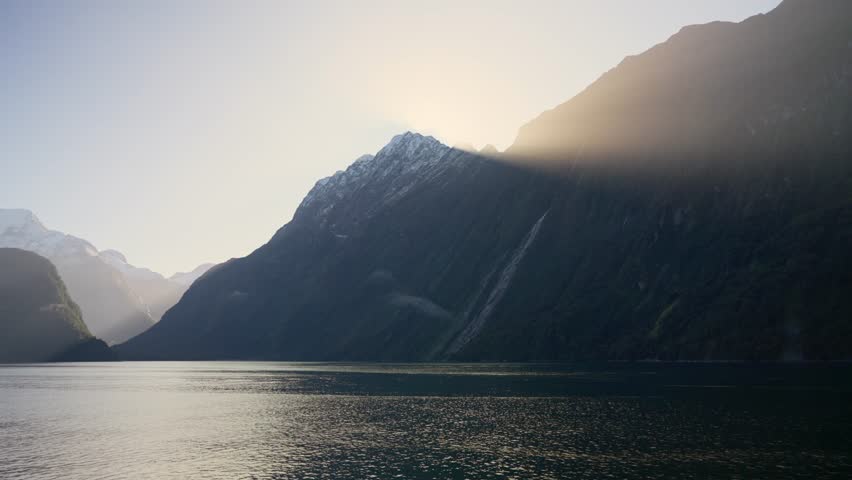 Epic orange warm sunlight shining through mountain peak in New Zealand. Fiords and steep cliffs by the water. Beautiful natural light and landscape in winter season in Fiordland in Milford Sound