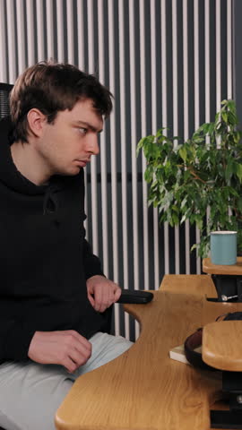 Having coffee, Vertical sipping, Hoodie drinking. Drinking coffee from blue cup, young man in black hoodie stands in front of vertical striped wall and green plant.