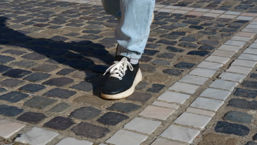 Person walking on cobblestone street. Casual walking in urban setting, black sneakers and light blue jeans stepping across gray and white cobblestone street during daytime travel journey