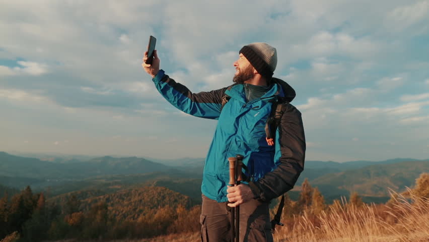 Solo traveler standing on a mountain slope tries to connect via mobile video call. Wind blows, signal is weak, nature surrounds him. Remote communication in wild highlands, solitude and technology ble