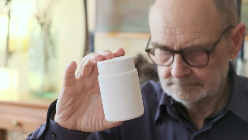 Man carefully looking at a small white medicine bottle in a cozy indoor room setting. He's wearing glasses, emphasizing an attentive and focused expression in a warm and domestic atmosphere.