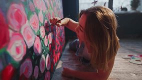 Young woman artist lying on the floor painting colorful roses on a large canvas in her art studio, illuminated by natural light from a nearby window - Powered by Shutterstock - Get 15% off with code: PIKWIZARD15