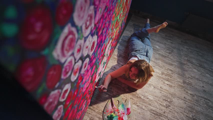 Talented female artist lying on the floor concentrates while painting colorful roses on a large canvas, showcasing her passion and dedication to art