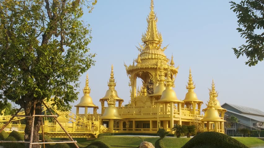 Golden Ganesha temple in Wat Rong Khun complex with ornate spires and traditional architecture surrounded by garden and pond under blue sky in sunny day, Chiang Rai, Thailand. White temple complex