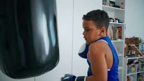 Determined young boy wearing blue boxing gloves, practicing powerful punches on black punching bag while developing athletic skills in home gym environment - Powered by Shutterstock - Get 15% off with code: PIKWIZARD15