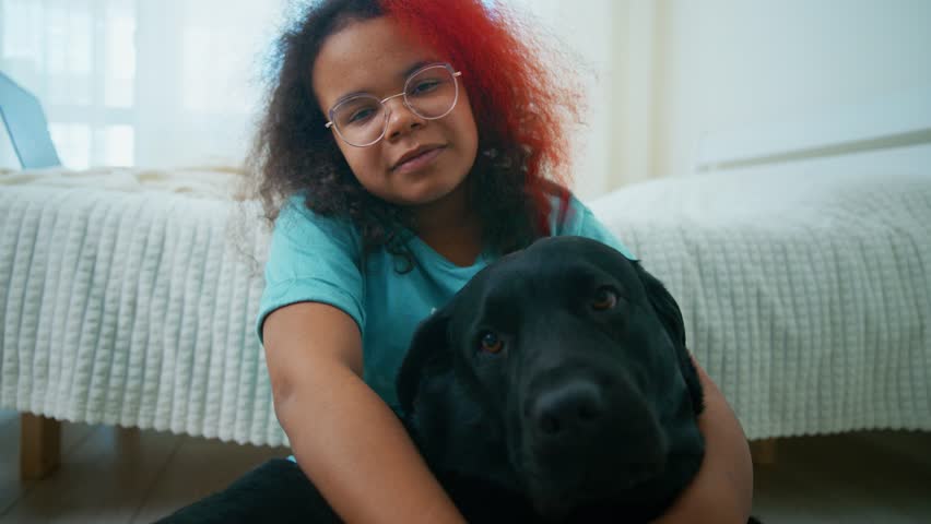 Young girl tenderly hugging black Labrador, sharing warm moment of affection while sitting on bedroom floor near bed, expressing deep bond between child and pet