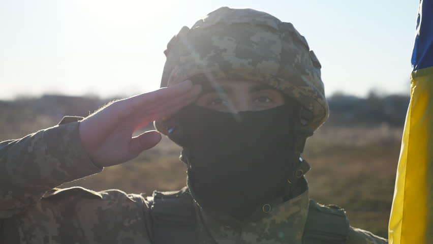 Male ukrainian army soldier in helmet and balaclava with national banner saluting against background of sunlight. Portrait of young military man with Ukraine flag at countryside. Invasion resistance