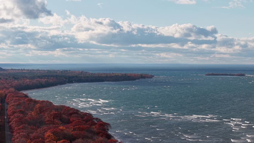 Aerial view of Lake Superior’s rugged coastline during fall, featuring vibrant red and orange forest meeting blue waters under scattered clouds