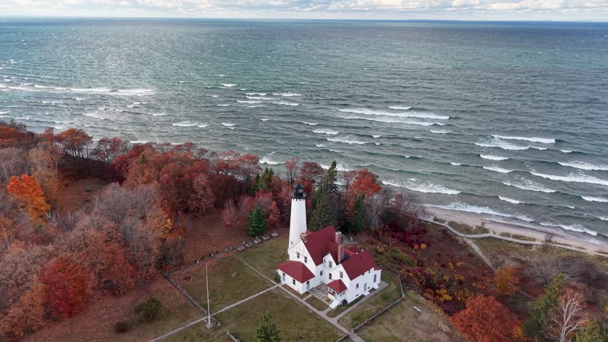 Aerial drone shot of Point Iroquois Lighthouse overlooking the choppy teal waters of Lake Superior, framed by brilliant fall foliage in Michigan’s Upper Peninsula