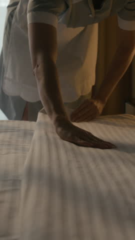 Vertical close up shot of hands of female housekeeper straightening striped bed sheet while cleaning hotel room