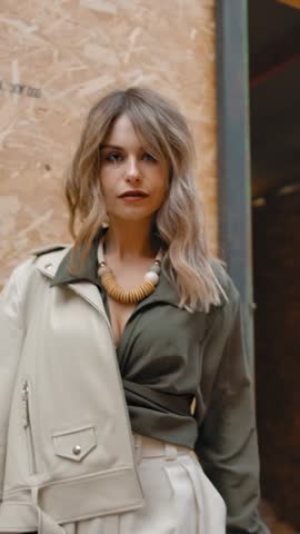 Beautiful woman in trendy elegant outfit stepping back in slow motion and looking at camera against open door of stall in stable on ranch. Vertical shot.