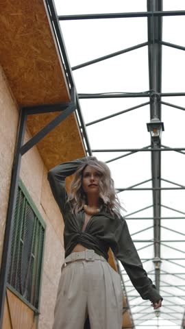 Low angle of elegant woman with hand on head shaking head in real time, then posing for camera in slow motion in corridor of barn on ranch. Vertical shot.